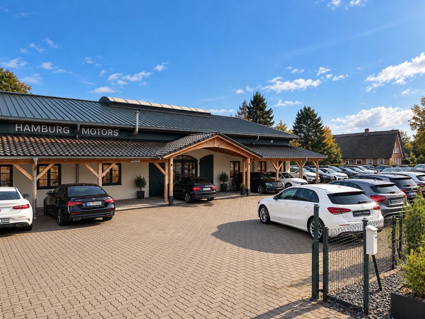 Eingang von Hamburg Motors am Tatenberger Deich: dunkelgrüne Holzfassade mit Schriftzug, Fahrzeuge unter dem Holzvordach und auf dem gepflasterten Hof